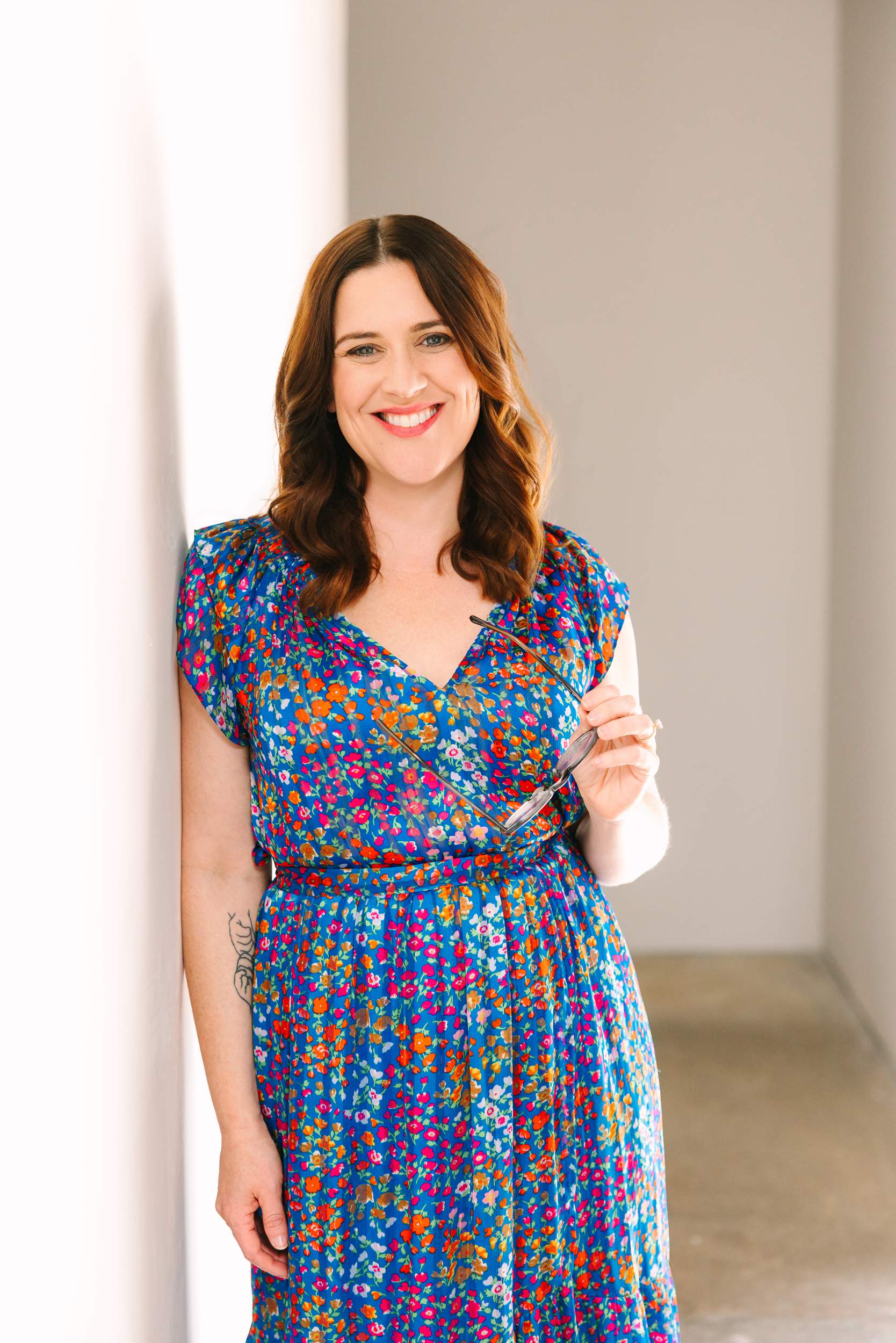 photo by Vallentyne Photography of A woman with wavy brown hair wearing a blue floral dress stands smiling against a white wall, holding eyeglasses in one hand. This softly lit, neutral scene is perfect for personal branding photography San Diego style.