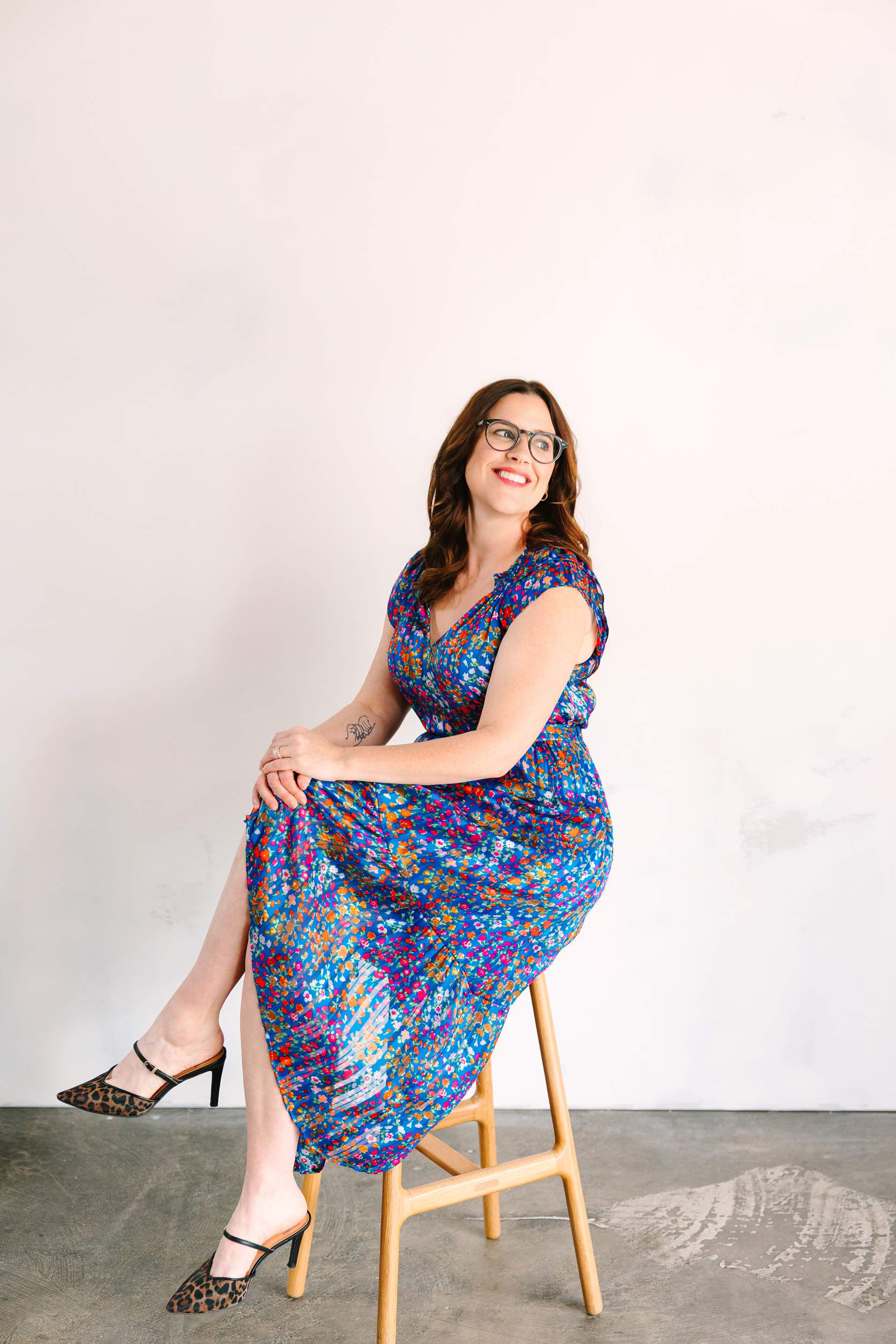 Woman in a colorful dress sits on a wooden stool, smiling and looking up, against a plain light background.