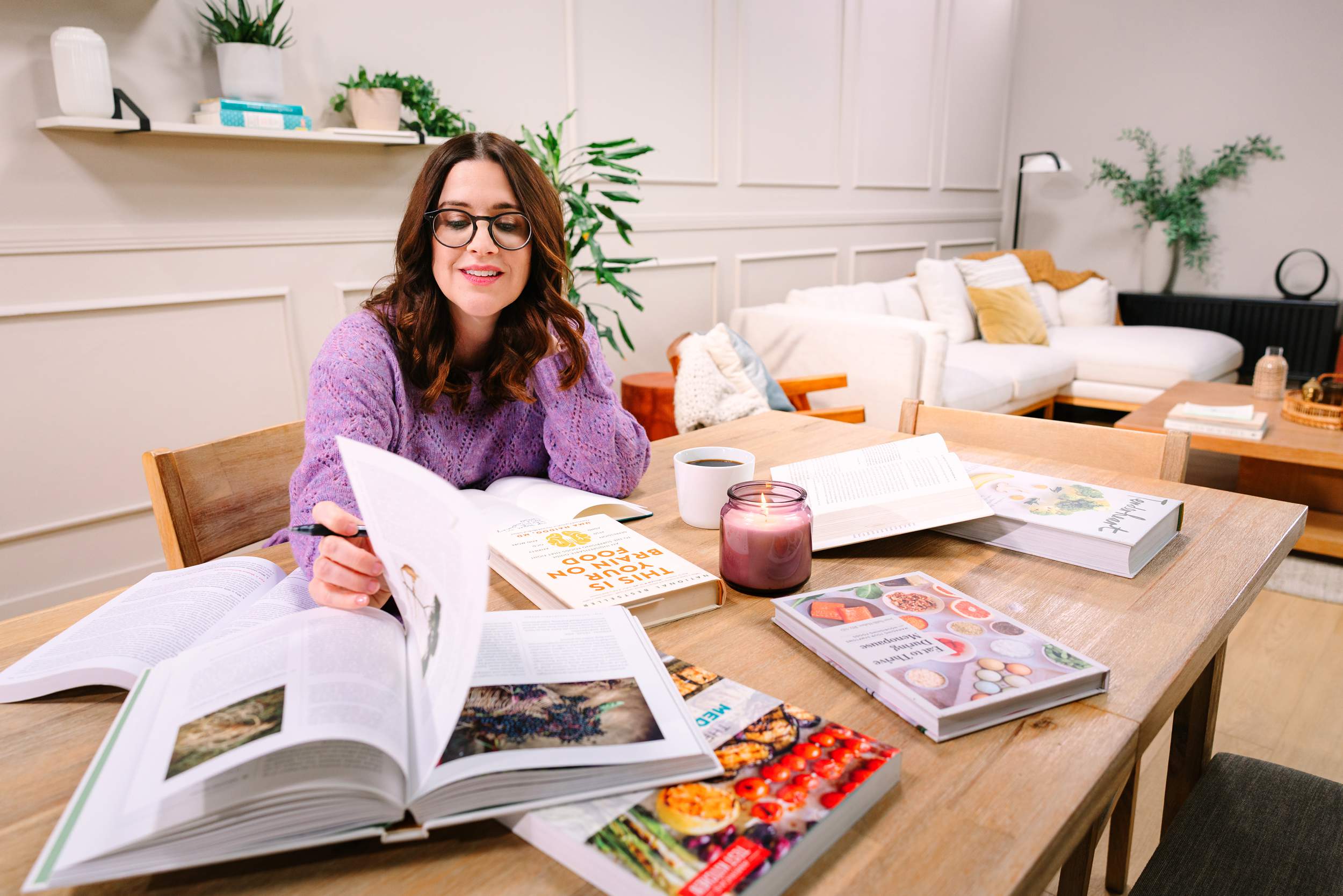 A woman in glasses studies at a table covered with books, a candle, and a coffee mug in a cozy living room.