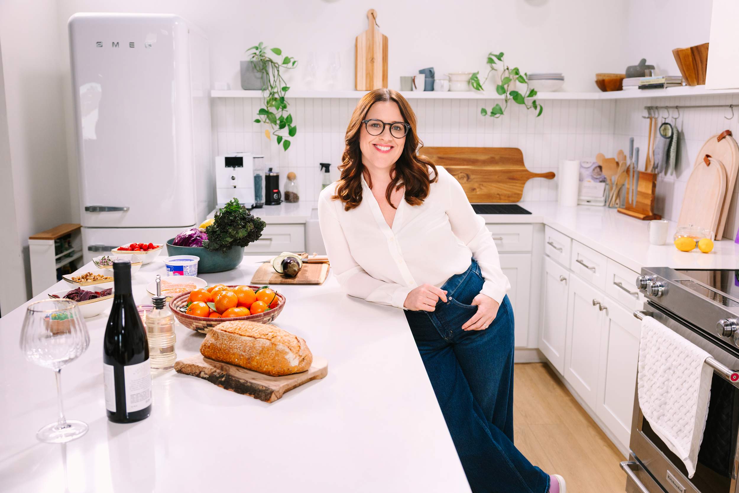 photo by Vallentyne Photography of A woman with glasses and a white blouse leans against a kitchen counter, surrounded by fresh vegetables, bread, and wine—captured by a personal branding photographer San Diego in a bright, modern kitchen.