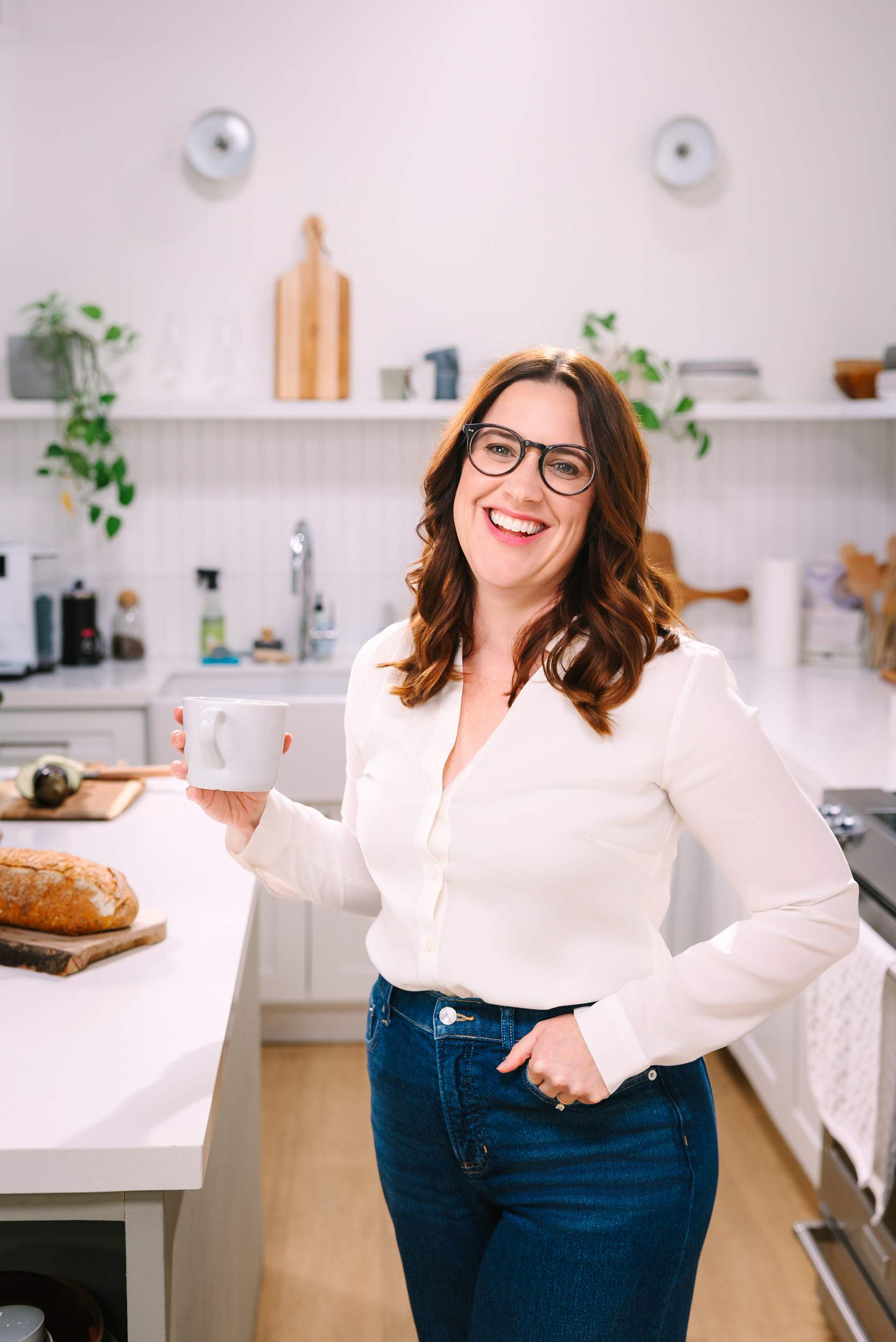 Woman with glasses in a white shirt smiles, holding a mug in a bright kitchen with bread on the counter.
