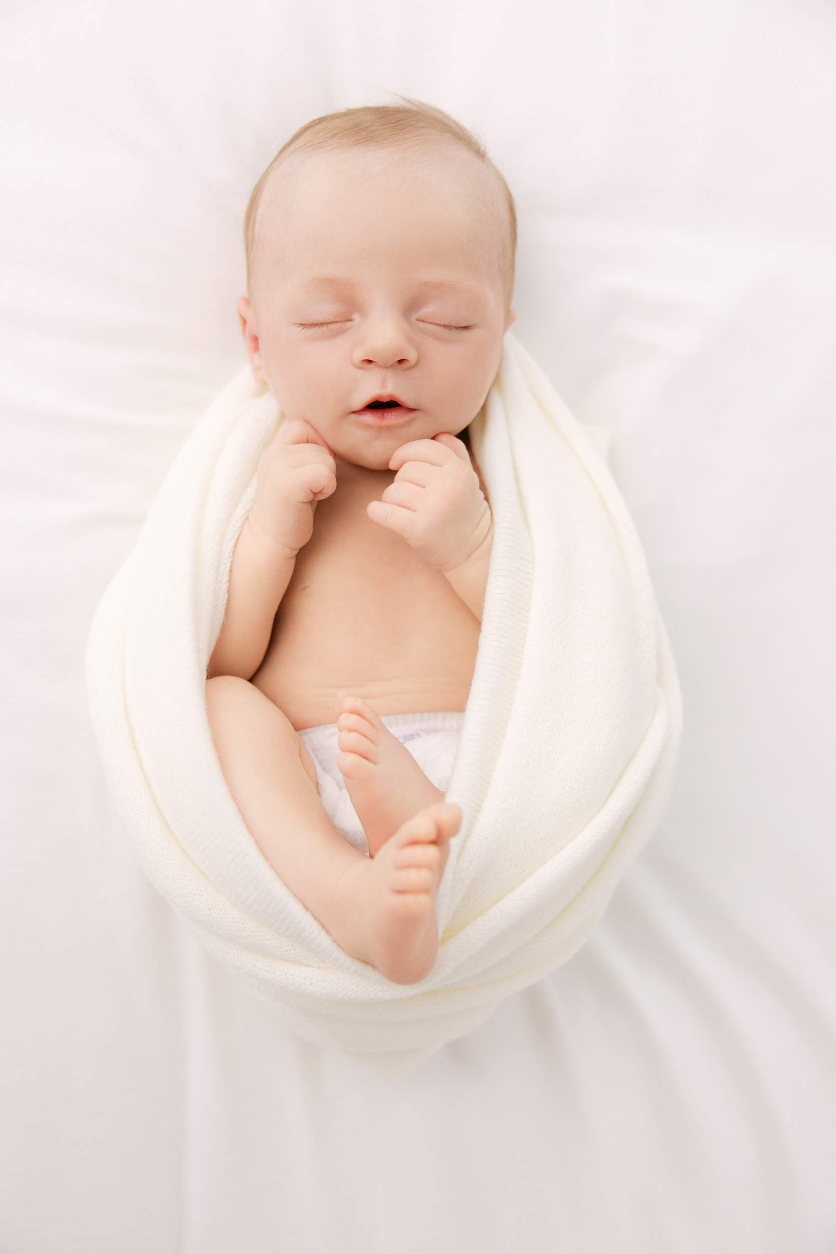 Newborn baby sleeping peacefully, wrapped in white, captured by newborn photography San Diego on a soft white surface.