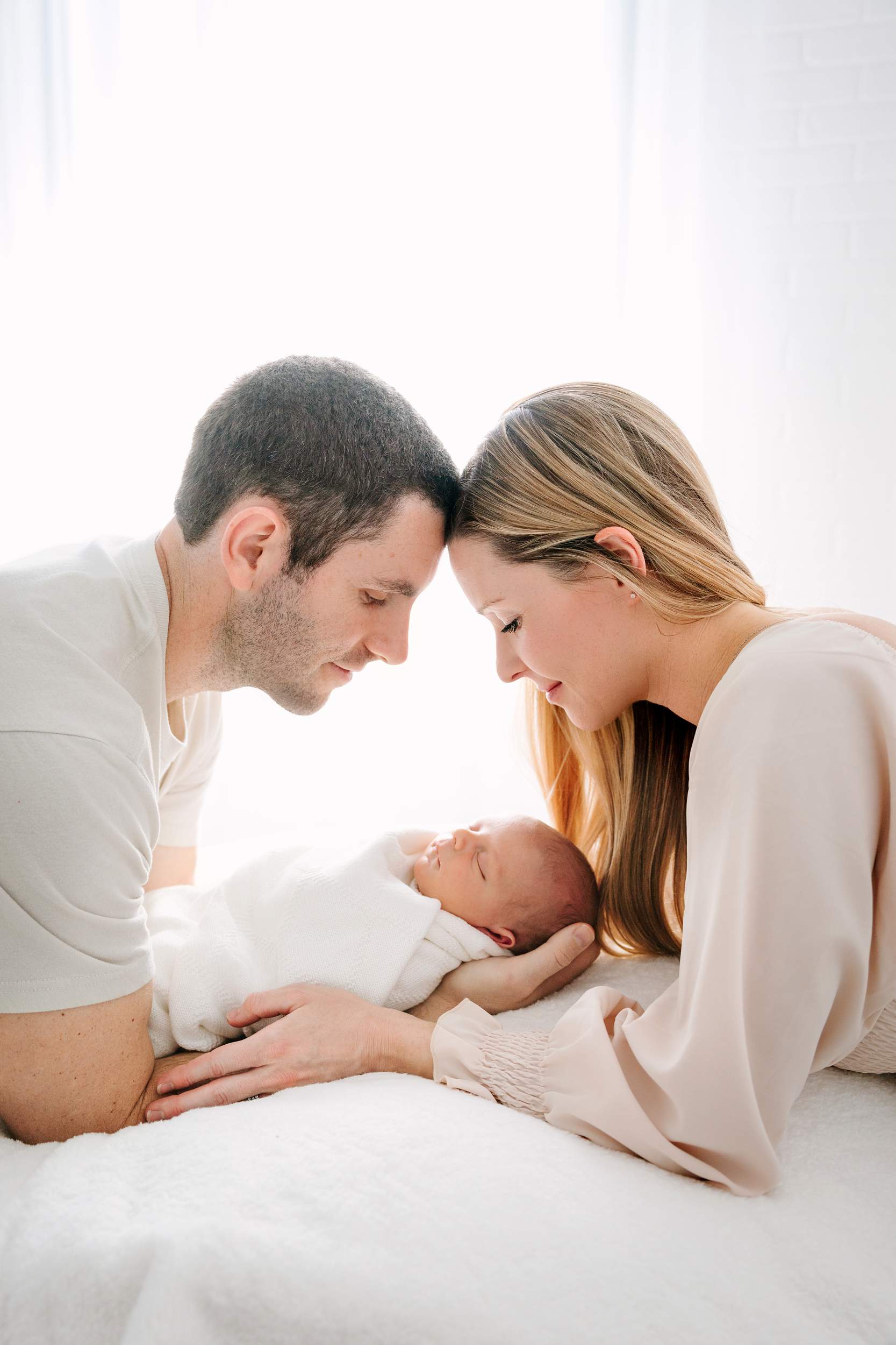A mother and father gently cradle their newborn baby, touching foreheads and smiling lovingly.