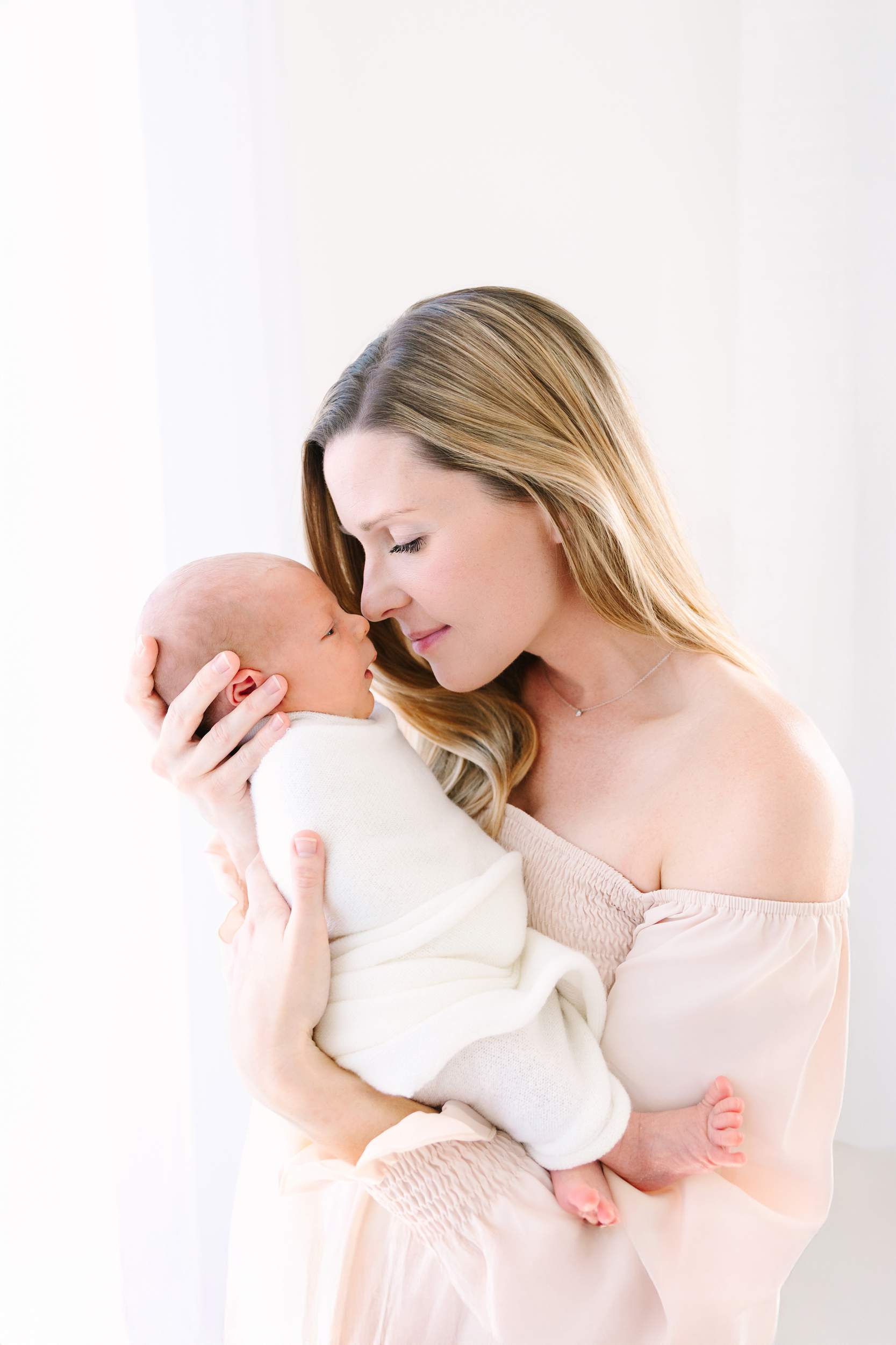 A woman gently holds a swaddled newborn close, touching noses in a bright, soft-lit room.