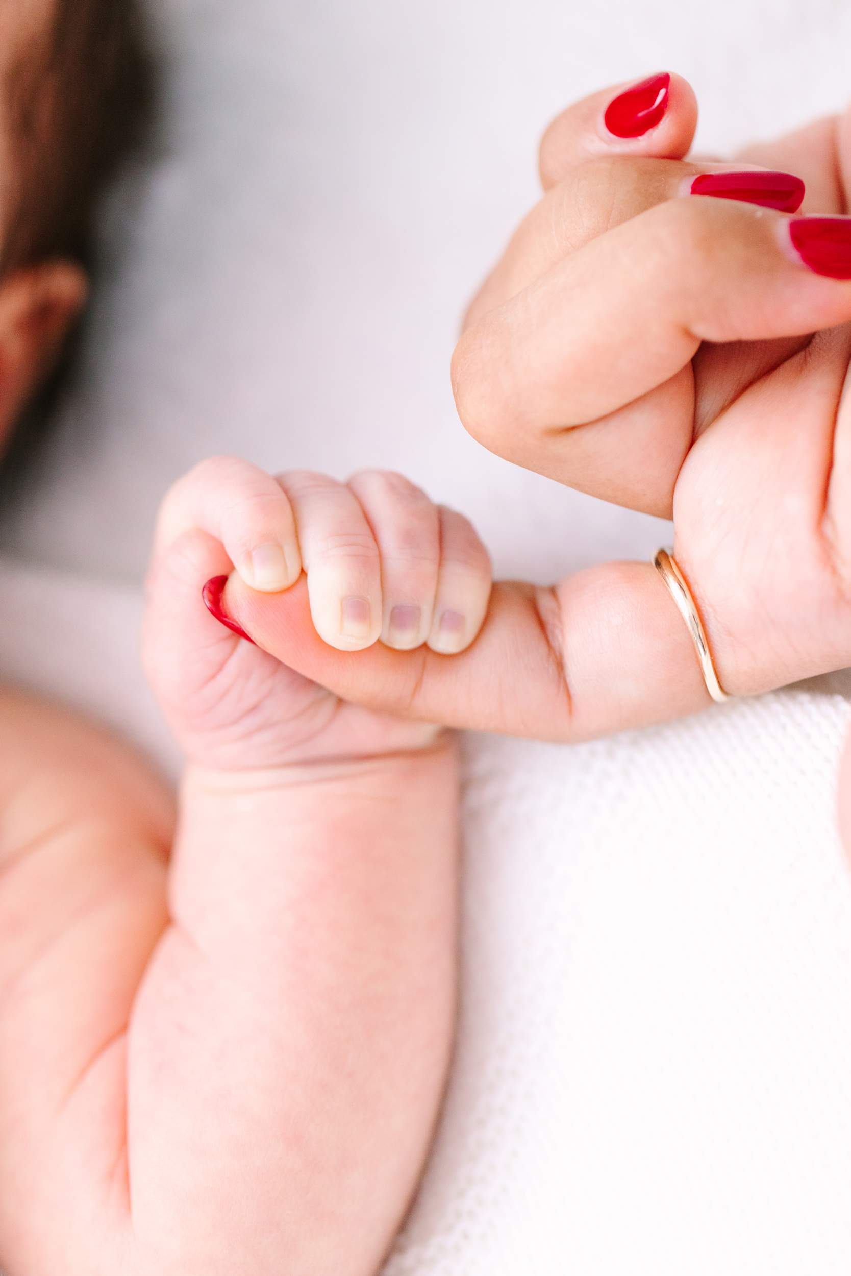 A baby gripping an adult’s finger, both with manicured nails and the adult wearing a gold ring.
