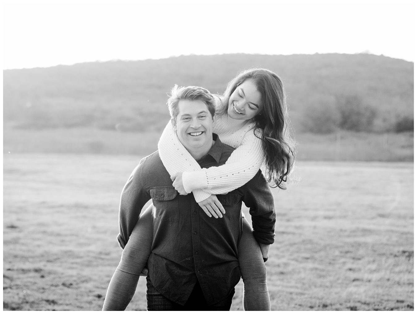 A smiling man gives a piggyback ride to a laughing woman in a grassy field with hills in the background.